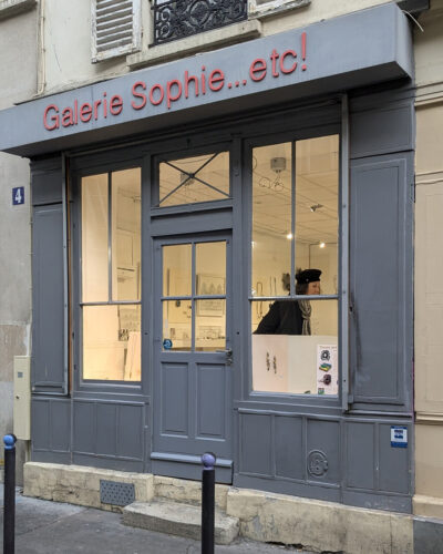 galerie Sophie...etc! view from the rue Gambey in Paris France. A gray exterior with red gallery name. A woman is visible inside looking at the jewelry